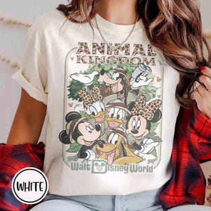 May include: Cream-colored t-shirt featuring a graphic of Disney characters, including Mickey, Minnie, Donald, Daisy, Goofy, and Pluto, with the text "ANIMAL KINGDOM" and "Walt Disney World". The characters are wearing safari hats.