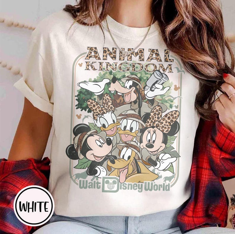 May include: Cream-colored t-shirt featuring a graphic of Disney characters, including Mickey, Minnie, Donald, Daisy, Goofy, and Pluto, with the text "ANIMAL KINGDOM" and "Walt Disney World". The characters are wearing safari hats.