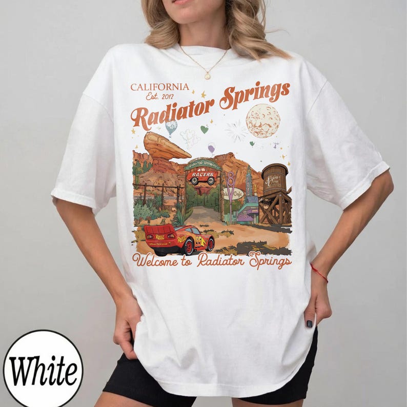 K&ouml;nnte beinhalten: Wei&szlig;es T-Shirt mit einem Radiator Springs-Motiv, mit dem Text "California Est. 2017" und "Welcome to Radiator Springs." Das Design zeigt ein rotes Rennauto und eine W&uuml;stenlandschaft.