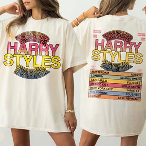 Pu&ograve; includere: Una t-shirt color avorio con una stampa grafica. La parte anteriore mostra il testo TOGETHER HARRY STYLES in rosa e giallo. Il retro presenta lo stesso testo sopra un elenco di citt&agrave; e nomi tra cui Amsterdam, London, Sao Paulo e Mexico City.