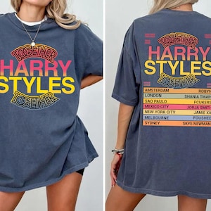Pu&ograve; includere: Una t-shirt blu scuro con una grafica sul davanti e sul retro. La parte anteriore mostra il testo "TOGETHER HARRY STYLES" in un design circolare. Il retro elenca citt&agrave; del tour e nomi tra cui "AMSTERDAM," "LONDON" e "HARRY STYLES."