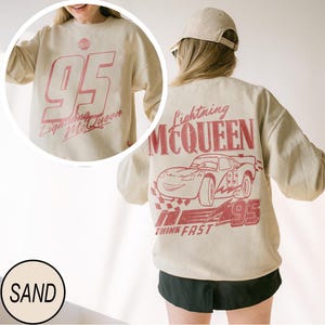 May include: Sand-colored sweatshirt featuring a large red "95" graphic with "Lightning McQueen" text on the front. The back displays a red Lightning McQueen car design with the text "Lightning McQueen" and "Think Fast".
