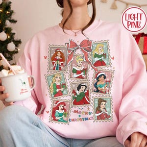 May include: Light pink sweatshirt featuring a collage of princess illustrations in postage stamp designs. A large pink bow is at the top, with the words "MERRY CHRISTMAS" below the stamps. The sweatshirt is a festive holiday theme.
