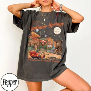 K&ouml;nnte beinhalten: Dunkelgraues T-Shirt mit einem Radiator Springs-Motiv aus dem Film Cars. Das Design enth&auml;lt den Text "CALIFORNIA Est. 2017 Radiator Springs" und "Welcome to Radiator Spring". Das Shirt hat kurze &Auml;rmel und eine lockere Passform.