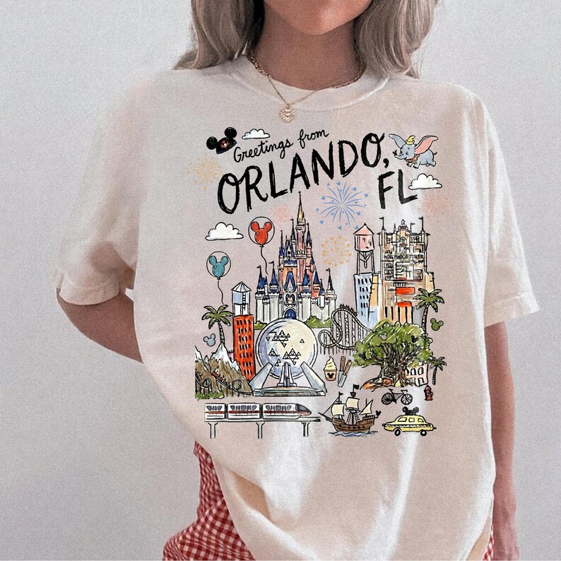 May include: Cream-colored t-shirt with a colorful illustration of Orlando, Florida landmarks. The design includes the text "Greetings from Orlando, FL" with images of a castle, roller coaster, and other attractions.