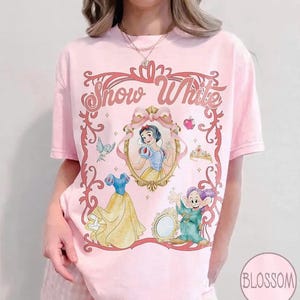 May include: Pink t-shirt featuring a Snow White design. The graphic includes an image of Snow White, a dress, a dwarf, and the words "Snow White" in a decorative frame. A crown, apple, and bird are also included.