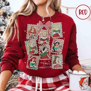 May include: A red sweatshirt featuring a collage of princess illustrations in postage stamp designs. A large pink bow is at the top, with the words "Merry Christmas" in the center. The sweatshirt is a solid red color.