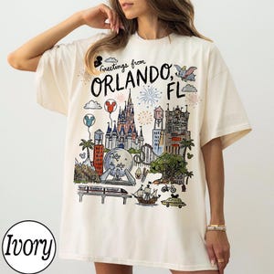 May include: Ivory-colored t-shirt with a colorful illustration of Orlando, FL landmarks, including a castle, roller coaster, and fireworks. The shirt features the text "Greetings from Orlando, FL" in a whimsical font. The shirt is a relaxed fit.