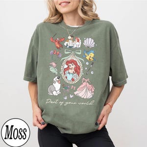Puede incluir: Camiseta verde oliva con una ilustraci&oacute;n enmarcada de Ariel de La Sirenita, rodeada de im&aacute;genes de dibujos animados. El texto "Part of your world" est&aacute; impreso debajo. La camiseta tiene cuello redondo y mangas cortas.