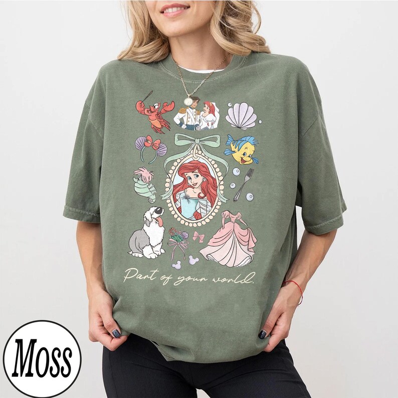 Puede incluir: Camiseta verde oliva con una ilustraci&oacute;n enmarcada de Ariel de La Sirenita, rodeada de im&aacute;genes de dibujos animados. El texto "Part of your world" est&aacute; impreso debajo. La camiseta tiene cuello redondo y mangas cortas.