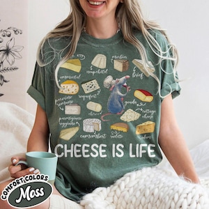 May include: Green t-shirt with a cheese-themed graphic. The design includes illustrations of various cheeses and a cartoon rat chef holding a wine glass. The text "CHEESE IS LIFE" is printed below the cheese illustrations.
