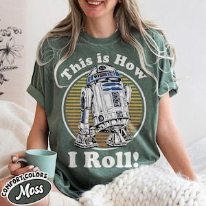 May include: A moss green t-shirt featuring a graphic of R2-D2 from Star Wars, with the text "This is How I Roll!" in a circular design. The shirt is a Comfort Colors brand.