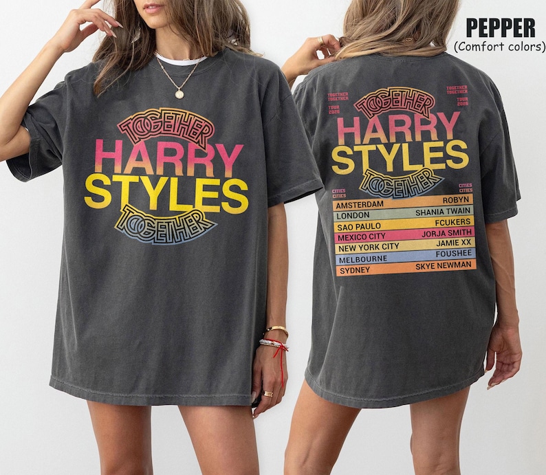 Pu&ograve; includere: Una t-shirt grigio antracite con una stampa grafica. La parte anteriore mostra il testo "TOGETHER HARRY STYLES" in rosa e giallo. Il retro mostra lo stesso testo sopra un elenco di citt&agrave; e nomi, tra cui "AMSTERDAM" e "LONDON".
