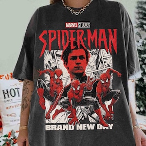 Puede incluir: Camiseta gris oscuro con un gr&aacute;fico de Spider-Man de Marvel Studios. El dise&ntilde;o incluye un retrato del actor y tres figuras de Spider-Man en poses de acci&oacute;n, con las palabras "BRAND NEW DAY" en la parte inferior.