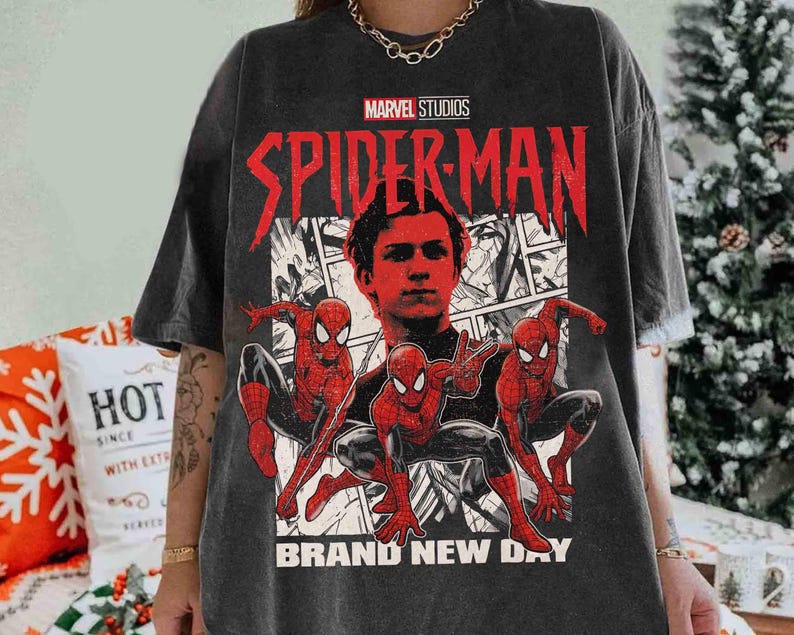Puede incluir: Camiseta gris oscuro con un gr&aacute;fico de Spider-Man de Marvel Studios. El dise&ntilde;o incluye un retrato del actor y tres figuras de Spider-Man en poses de acci&oacute;n, con las palabras "BRAND NEW DAY" en la parte inferior.
