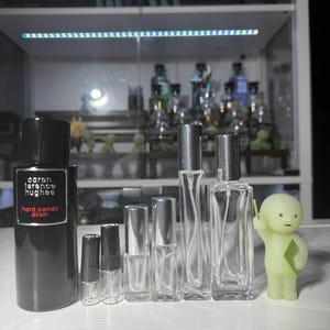 May include: A collection of perfume bottles and a small figurine. The largest bottle is black with white text that reads "Aaron Terence Hughes Hard Candy Elixir." Several clear glass bottles of varying sizes are arranged in a row. A small, green figurine stands to the right.