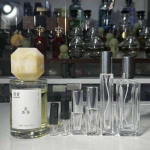 May include: A collection of clear glass perfume bottles of various sizes, some with silver sprayers and black caps. One bottle has a large, light-colored, geometric stopper. The bottles are arranged in a row, with a shelf of other perfume bottles in the background.