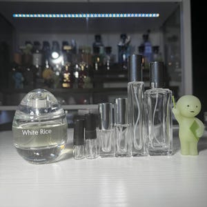 May include: A collection of perfume bottles and a small figurine. The bottles vary in size and shape, including an egg-shaped bottle with "White Rice" printed on it. The bottles are clear with black caps.