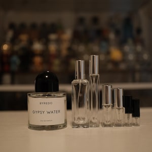 May include: A collection of perfume bottles and travel-sized atomizers. The main bottle is labeled "BYREDO GYPSY WATER" with a black cap. Several clear glass spray bottles and small vials are arranged beside it, suggesting fragrance samples or travel sizes.