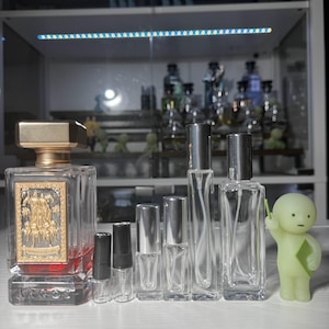 May include: A collection of perfume bottles and atomizers of varying sizes. The largest bottle has a gold-colored cap and a decorative front. A small, green figurine stands to the right. The background shows a cabinet with more perfume bottles.