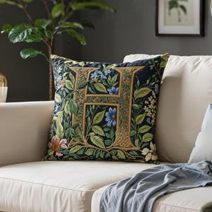 Op de afbeelding: Een decoratief kussen met de gouden letter "H", omringd door bloemmotieven in groen, blauw en roze. Het ligt op een crèmekleurige bank met een blauwe deken. De kamer heeft een gezellige, uitnodigende sfeer.