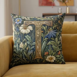 Op de afbeelding: Een decoratief sierkussen met een bloemenontwerp en de letter "T" in het midden. Het kussen heeft een donkerblauwe achtergrond met witte, blauwe en oranje bloemen en groene bladeren. Het kussen ligt op een gele bank.