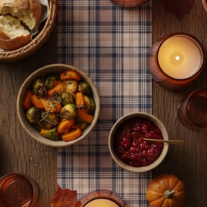 May include: A table set for a meal, featuring lit candles in amber glass holders, bowls of roasted vegetables, cranberry sauce, and rolls. A plaid runner and small pumpkin enhance the autumn theme. The scene is set on a wooden table.