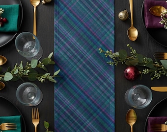 May include: A festive table setting with a blue, green, and purple plaid table runner. The table is set with black plates, gold cutlery, clear glasses, and green and purple napkins. Decorative ornaments and greenery complete the holiday theme.
