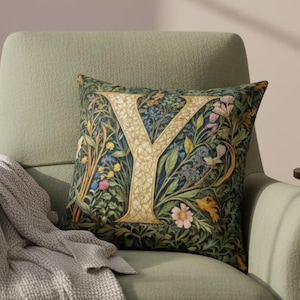 Op de afbeelding: Een decoratief sierkussen met een bloemenpatroon en de gouden letter "Y". Het kussen ligt op een groene fauteuil met een grijze deken. Een kleine tafel heeft een vaas met bloemen en boeken.