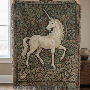 May include: A tapestry throw blanket featuring a white unicorn against a dark green background with floral and tree motifs. The blanket has a fringed edge and is held up in front of a person.