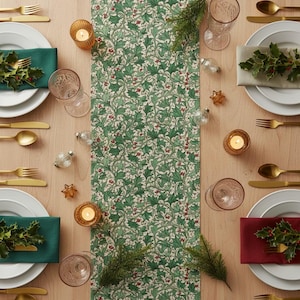 Puede incluir: Una mesa festiva con platos blancos, cubiertos dorados y servilletas de colores. Un camino de mesa verde con estampado de acebo recorre el centro. Vasos transparentes, pequeños adornos y velas encendidas completan el tema navideño.