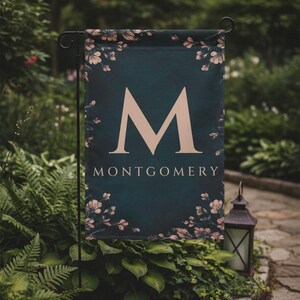 May include: A dark teal garden flag with the name "Montgomery" and a large letter "M" in a light beige colour. The flag is decorated with floral accents and is displayed outdoors in a garden setting.