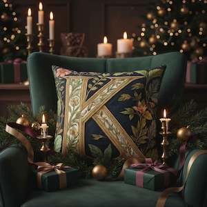 Op de afbeelding: Een decoratief kussen met een grote gouden en crèmekleurige letter "K", omringd door bloemmotieven op een marineblauwe achtergrond. Het kussen ligt op een groene fauteuil, versierd met kaarsen, ballen en ingepakte cadeaus.