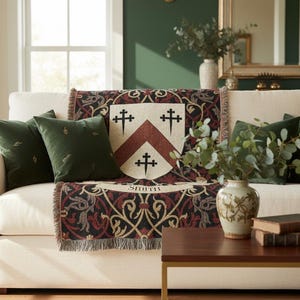 May include: A cream-colored sofa with a decorative throw blanket featuring a family crest design. The blanket has a cream background with a burgundy and black pattern. Two green velvet pillows and a vase of greenery complete the scene.