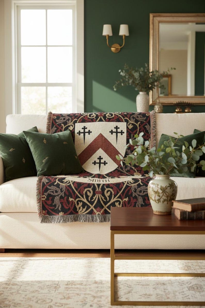 May include: A cream-colored sofa with a decorative throw blanket featuring a family crest design. The blanket has a cream background with a burgundy and black pattern. Two green velvet pillows and a vase of greenery complete the scene.