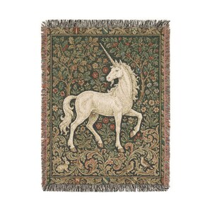 May include: A woven tapestry throw blanket featuring a white unicorn with a long horn, set against a backdrop of a forest with trees, flowers, and foliage in shades of green, red, and brown. The blanket has a fringed edge.