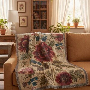 May include: A floral throw blanket with a beige background and large burgundy flowers with green leaves. The blanket is draped over a brown sofa in a living room setting. The room has a wooden cabinet, framed botanical prints, and a white lamp.