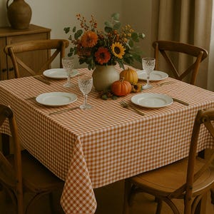 May include: A dining table set for a meal, featuring a tablecloth with a brown and white gingham pattern. The table is set with white plates, crystal glasses, and gold-colored cutlery. A vase of flowers and pumpkins complete the autumn-themed decor.