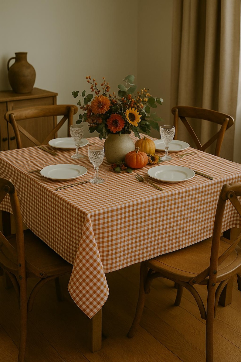 May include: A dining table set for a meal, featuring a tablecloth with a brown and white gingham pattern. The table is set with white plates, crystal glasses, and gold-colored cutlery. A vase of flowers and pumpkins complete the autumn-themed decor.