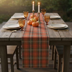 Puede incluir: Una mesa rústica de madera preparada para una reunión de otoño. La mesa está adornada con un camino de mesa a cuadros en tonos naranja, verde y blanco. Platos blancos, cubiertos dorados y vasos color ámbar están colocados. Calabazas en miniatura y velas encendidas completan la escena otoñal.