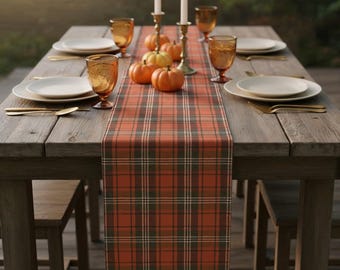 May include: A rustic wooden table set for a fall gathering. The table is adorned with a plaid table runner in shades of orange, green, and white. White plates, gold cutlery, and amber-colored glasses are set for each place setting. Miniature pumpkins and lit candles complete the autumnal scene.