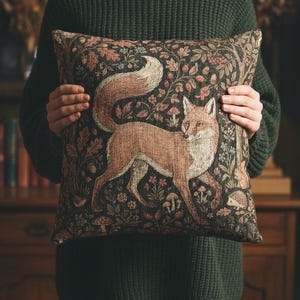 May include: Decorative throw pillow featuring a detailed illustration of a fox in a forest setting. The pillow has a dark background with floral and mushroom accents. The fox is depicted in shades of brown and white.