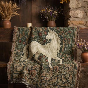 May include: A tapestry throw blanket featuring a white unicorn design on a dark green background with floral and tree patterns. The blanket has a fringed edge and is draped over a wooden bench. Decorative dried flowers and wheat are in the background.