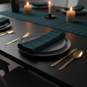 May include: A table setting featuring a dark teal and black plaid napkin on a white plate. Gold-coloured cutlery, including a spoon, fork, and knife, are arranged beside the plate. Lit candles and a plaid table runner complete the elegant setup.