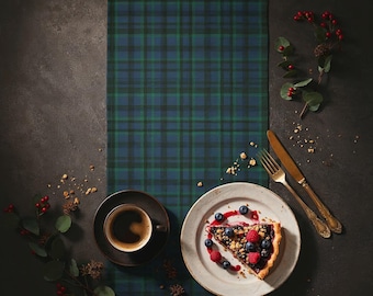 May include: A slice of berry tart on a white plate, with a cup of coffee and a fork and knife. The scene is set on a blue and green tartan table runner. Berries and greenery add colour to the dark background.