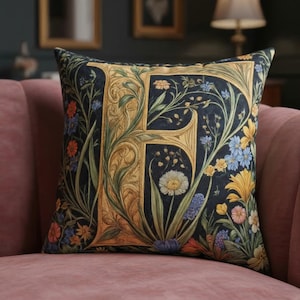 Op de afbeelding: Vierkant decoratief kussen met een grote, sierlijke gouden letter "F" op een donkerblauwe achtergrond. Het kussen is versierd met kleurrijke bloemen- en bladpatronen in geel, blauw en groen. Het kussen ligt op een roze bank. Afmetingen: ongeveer 40 cm x 40 cm.