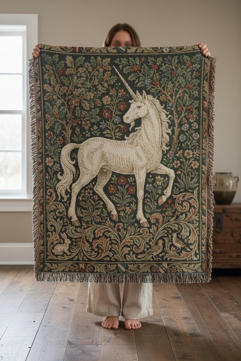 May include: A tapestry throw blanket featuring a white unicorn against a dark green background with floral and tree motifs. The blanket has a fringed edge and is held up in front of a person.
