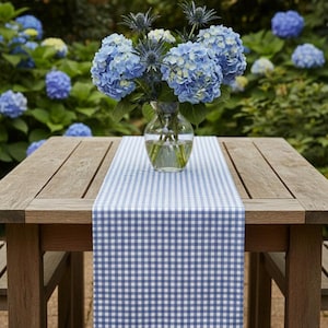 Op de afbeelding: Een houten tuintafel met een blauw-witte geruite tafelloper. Een glazen vaas toont blauwe hortensia's en distels. De achtergrond toont een groene tuin met blauwe hortensiastruiken.