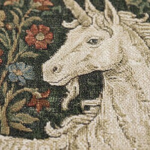 May include: Close-up of a tapestry featuring a unicorn with a long, spiraled horn. The unicorn is cream-colored, set against a dark green background with red, blue, and green floral elements. The tapestry has a textured appearance.