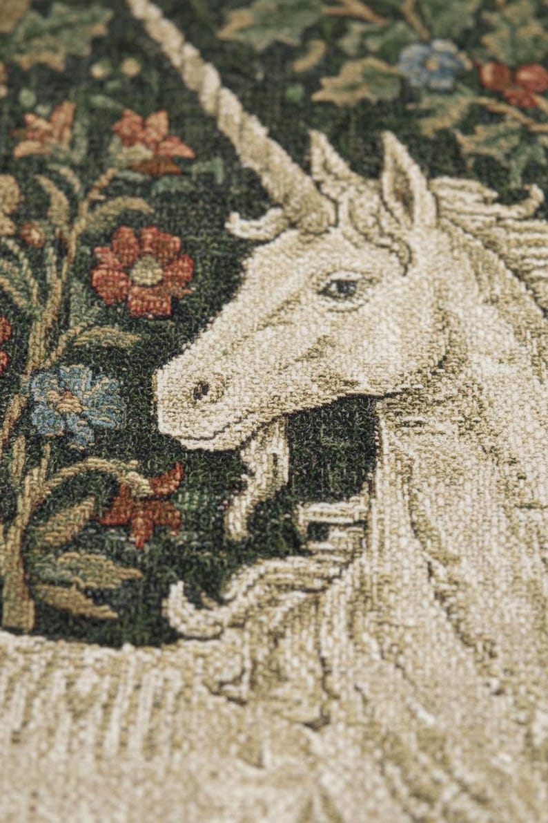 May include: Close-up of a tapestry featuring a unicorn with a long, spiraled horn. The unicorn is cream-colored, set against a dark green background with red, blue, and green floral elements. The tapestry has a textured appearance.